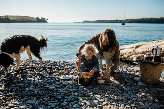 A woman and toddler stirring a pot on a pebbly beach.