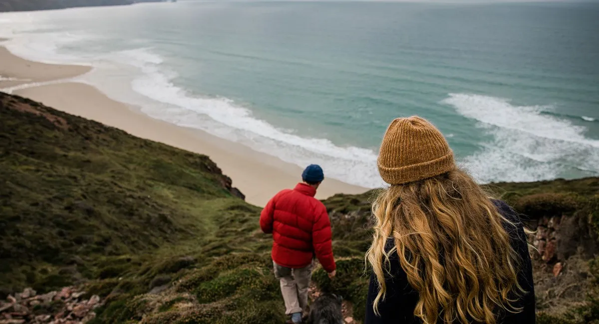 A couple walking a dog on a cliff overlooking a beach.