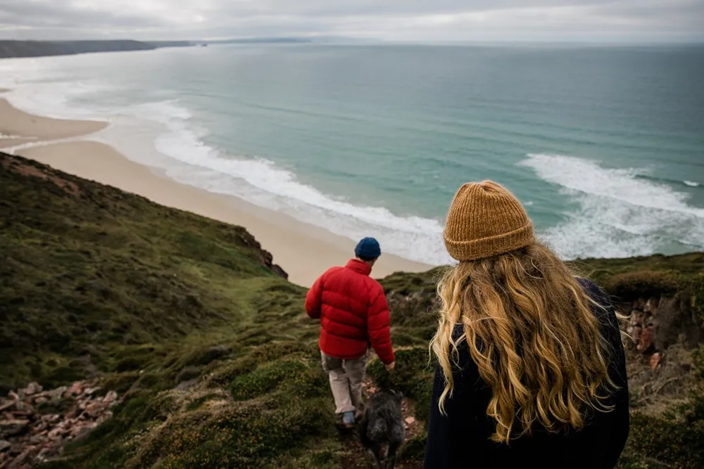 A couple walking a dog on a cliff overlooking a beach.
